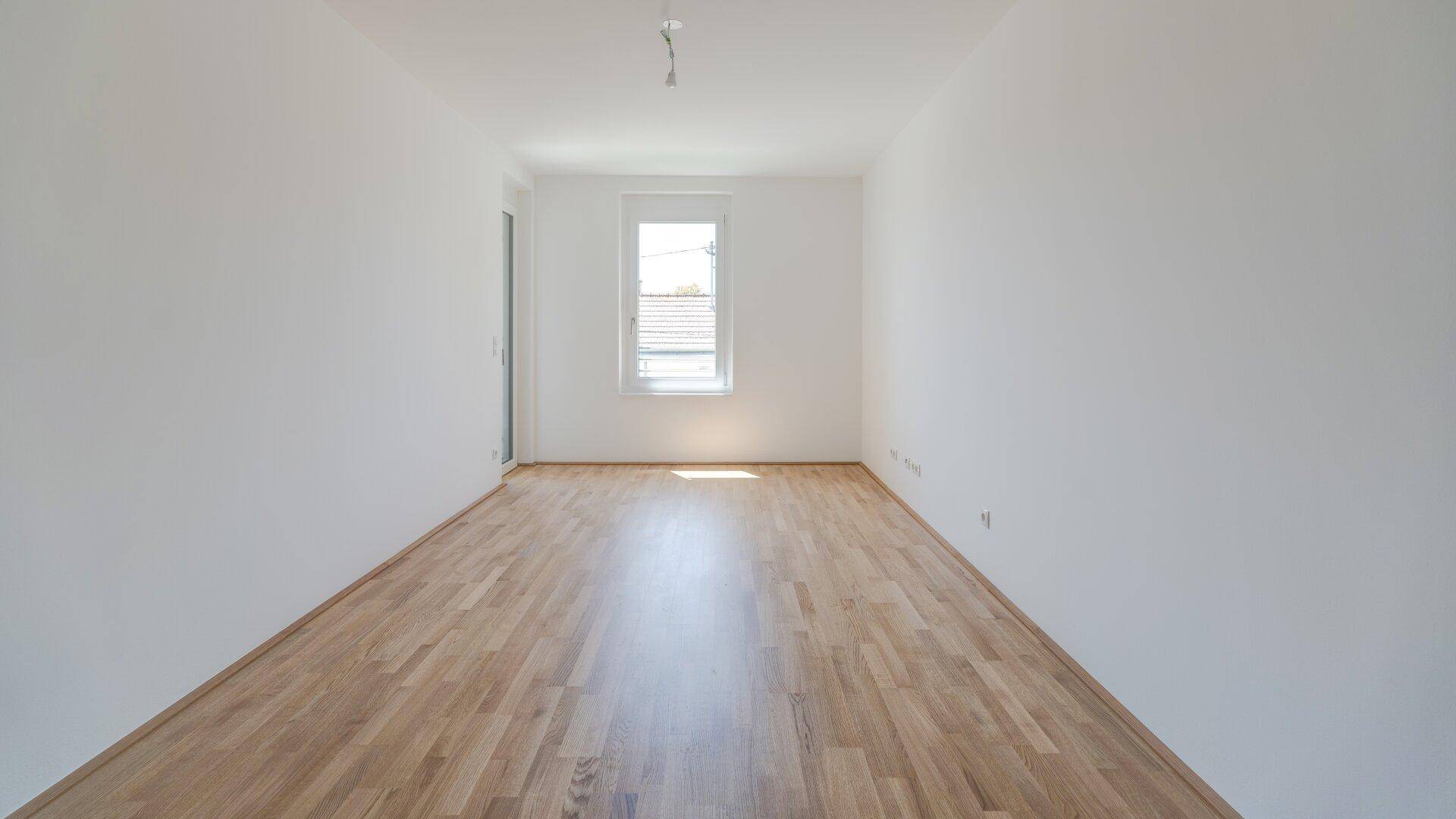 Langer, heller Raum mit Holzparkettboden und einem Fenster am Ende, bereit zur Einrichtung.