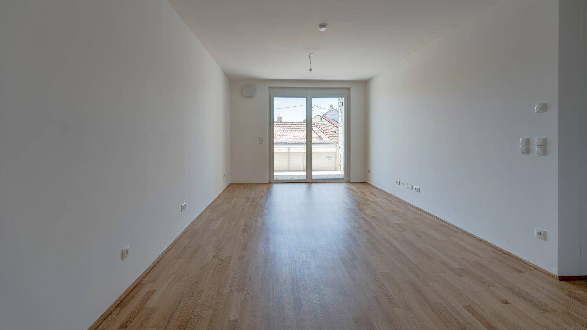 Geräumiger, heller Raum mit Holzparkettboden und Zugang zu einem Balkon durch eine große Schiebetür.