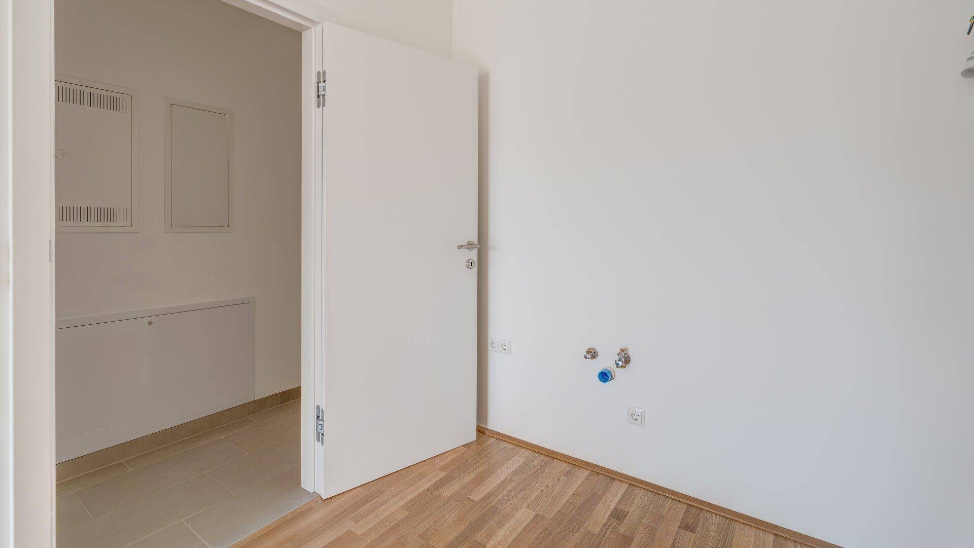 Leerer Raum mit Holzparkettboden und Anschlüssen für Waschmaschine oder andere Geräte.