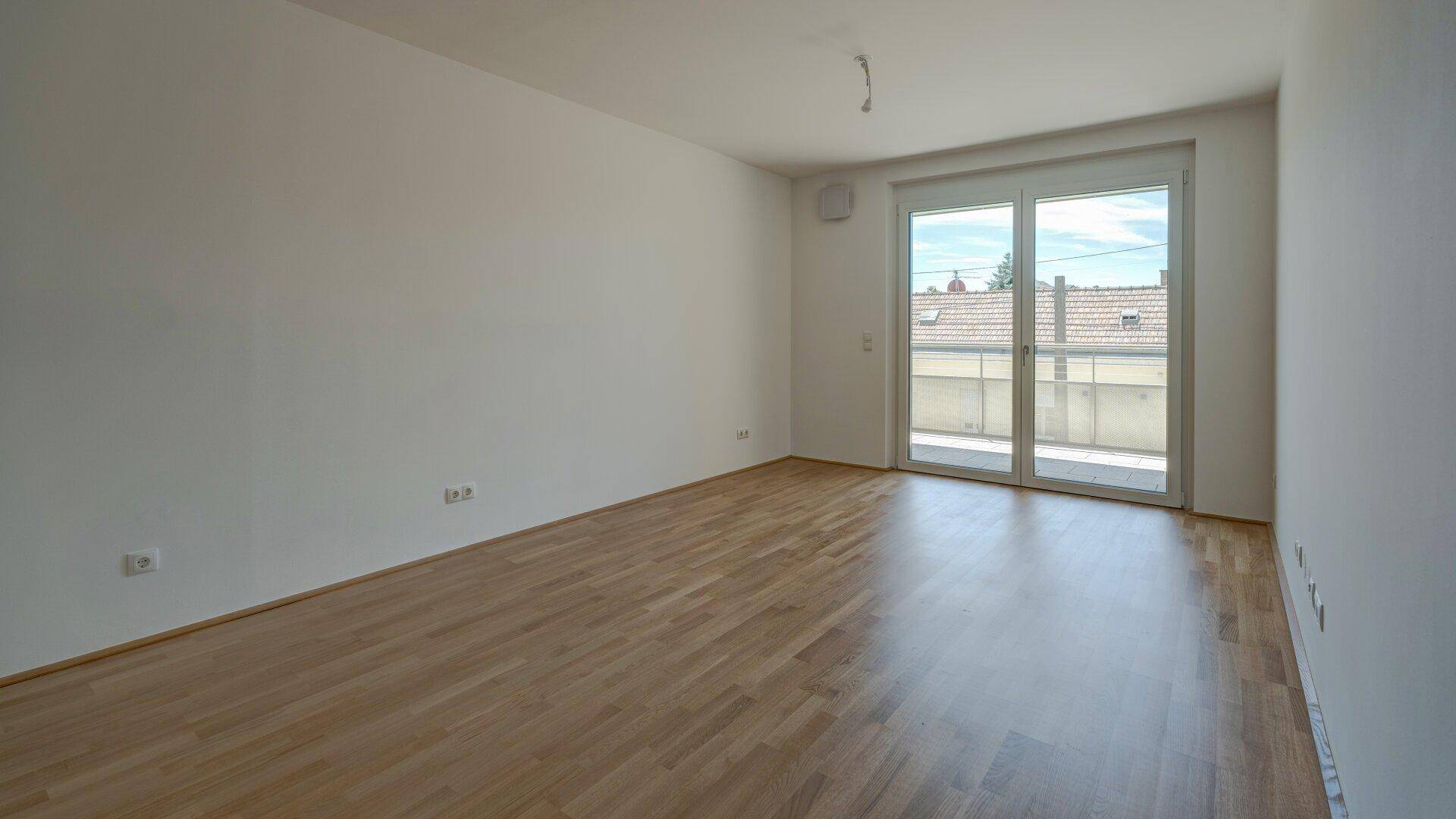 Geräumiger, heller Raum mit Holzparkettboden und großen Fenstern, die auf einen Balkon führen.