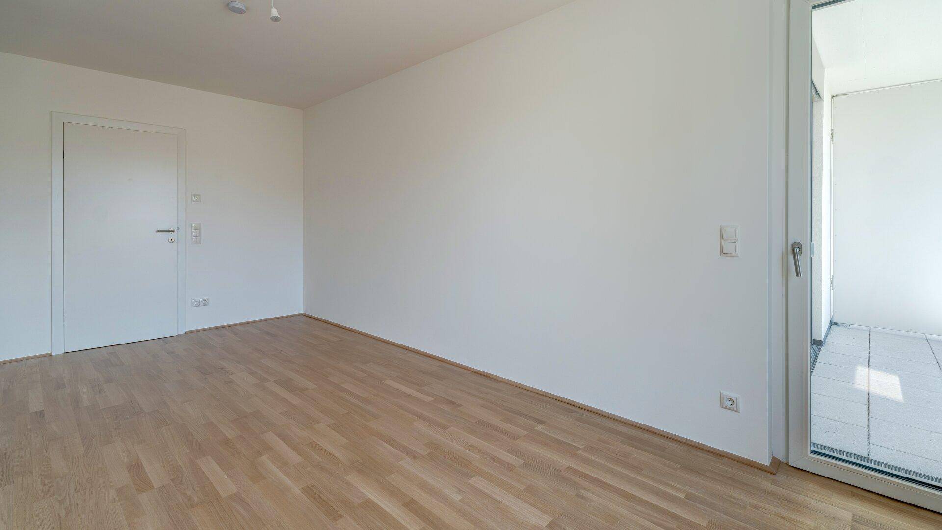 Heller Raum mit Holzparkettboden und einer Tür, die zu einem Balkon führt.