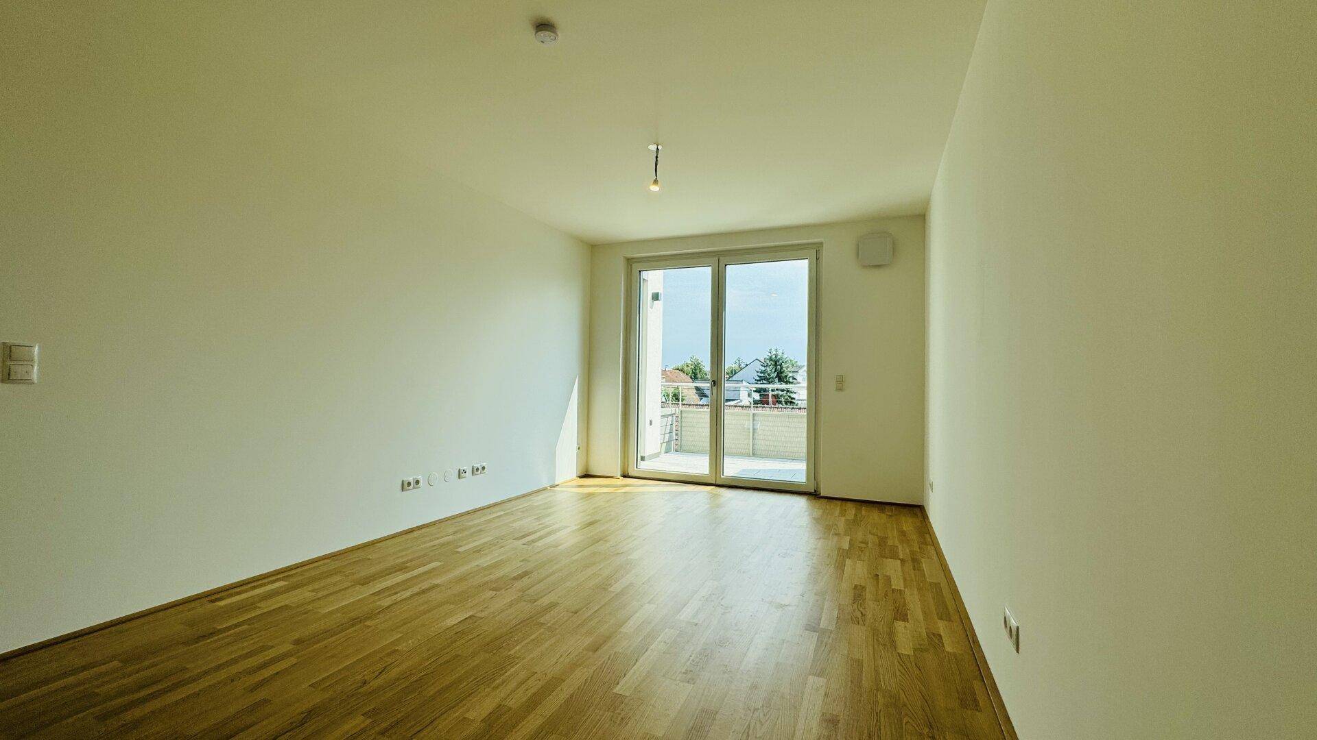 Heller Raum mit Holzboden und großer Schiebetür, die Zugang zu einem sonnigen Balkon bietet.
