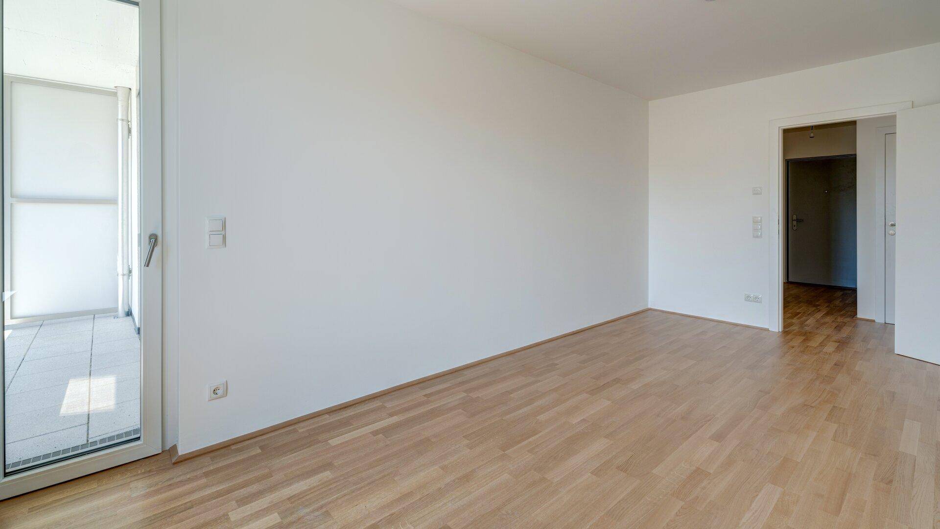 Geräumiges, helles Zimmer mit Holzboden und Balkonzugang, mit Blick auf die Umgebung.