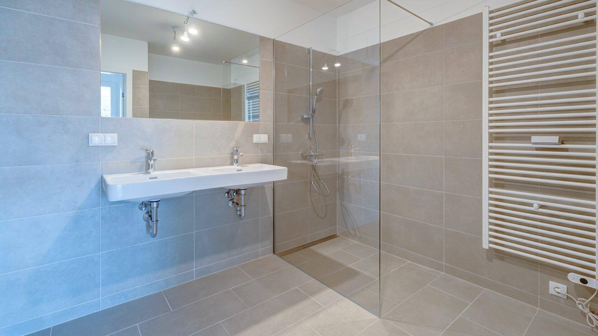 Modernes Badezimmer mit grauen Fliesen, Doppelwaschbecken, begehbarer Dusche und Handtuchheizkörper.