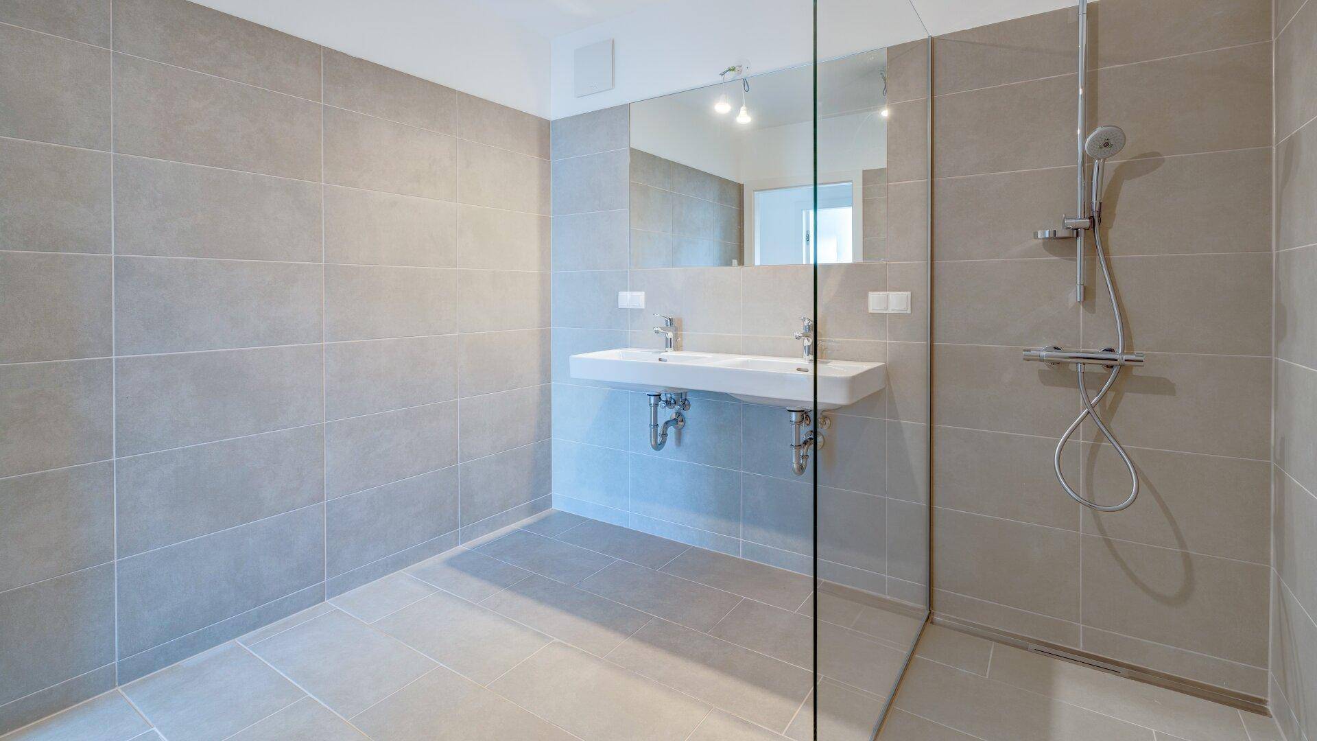 Geräumiges Badezimmer mit grauen Fliesen, Doppelwaschbecken und großer begehbarer Dusche.