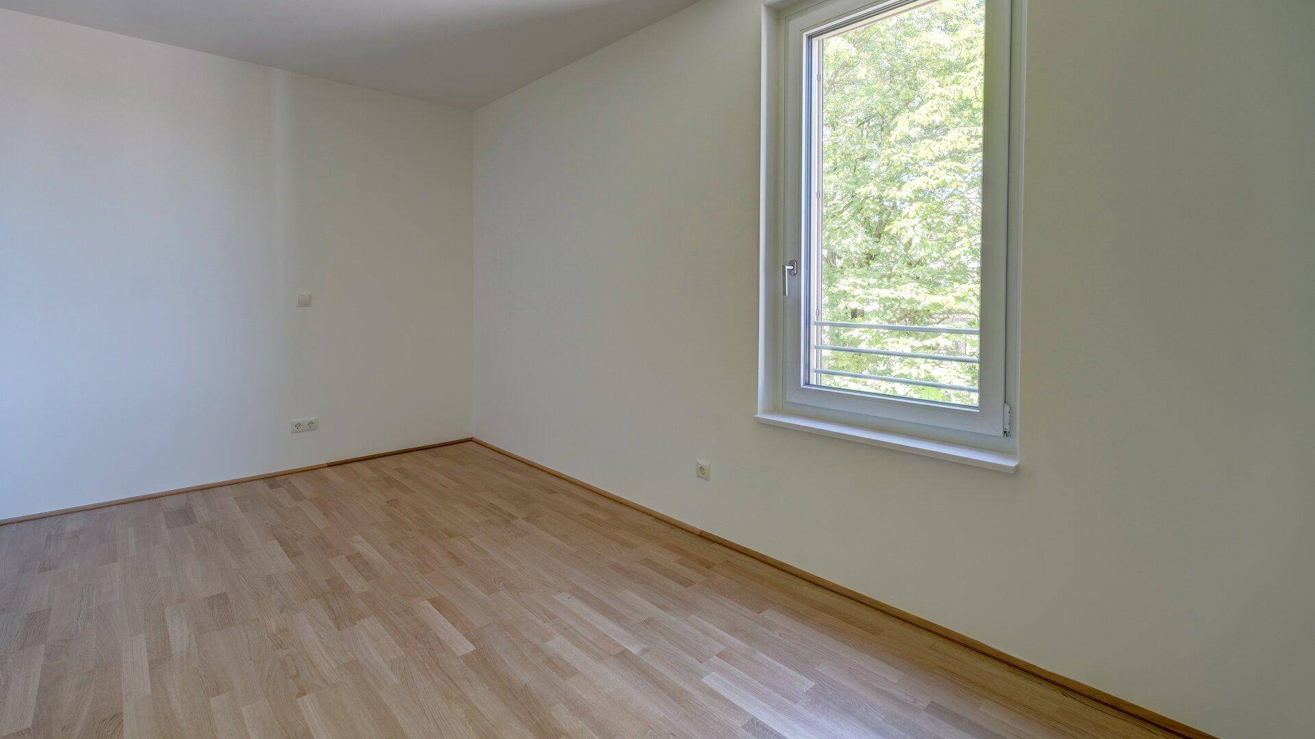 Helles, leeres Zimmer mit Holzparkettboden und einem großen Fenster mit Blick ins Grüne.