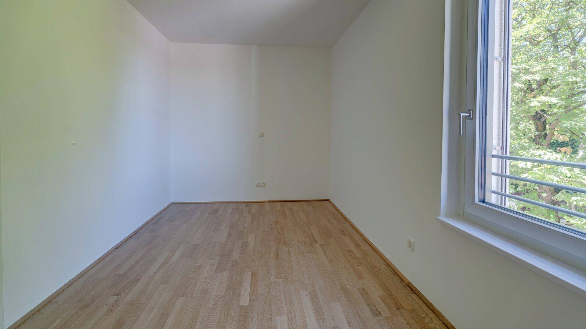 Lichtdurchflutetes Zimmer mit hellem Holzparkett und einem Fenster, das viel Tageslicht bietet.