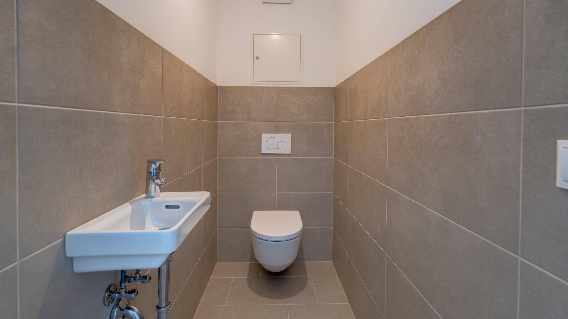 Modernes Gäste-WC mit grauen Fliesen, wandhängendem WC und einem kleinen Waschbecken.