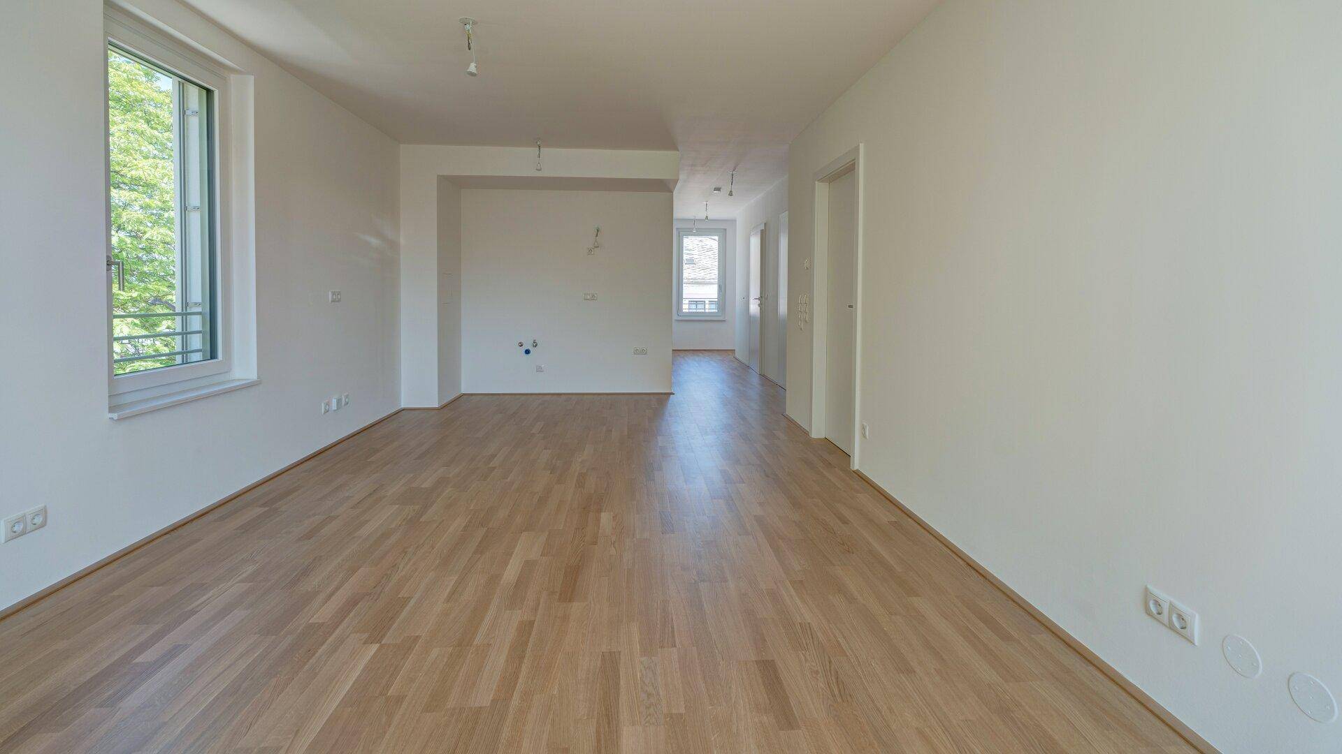 Langer, heller Wohnbereich mit Holzparkettboden und Anschlüssen für eine offene Küche.
