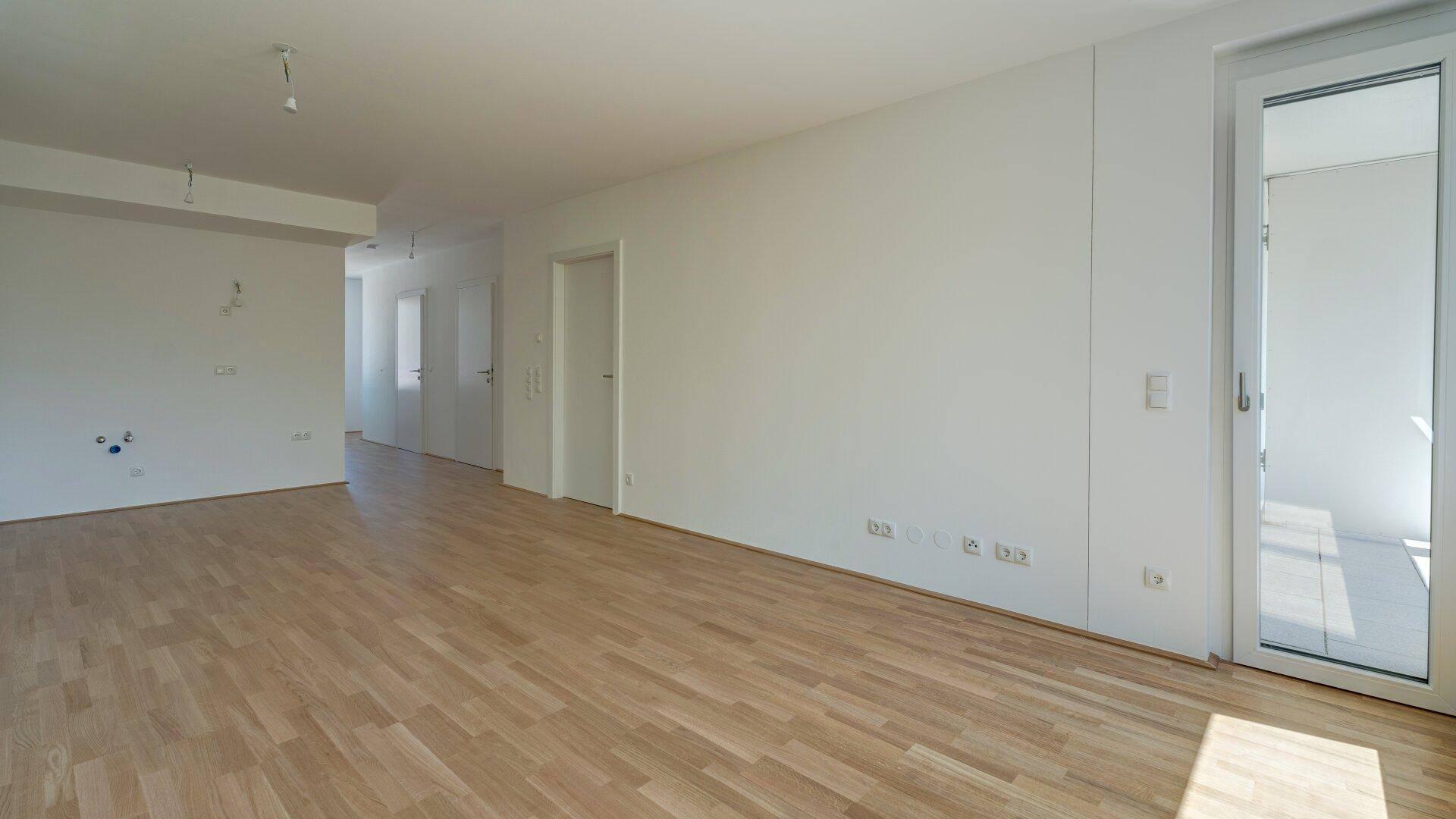 Heller Wohnbereich mit Holzparkettboden und Zugang zu einem Außenbereich durch eine Glastür.