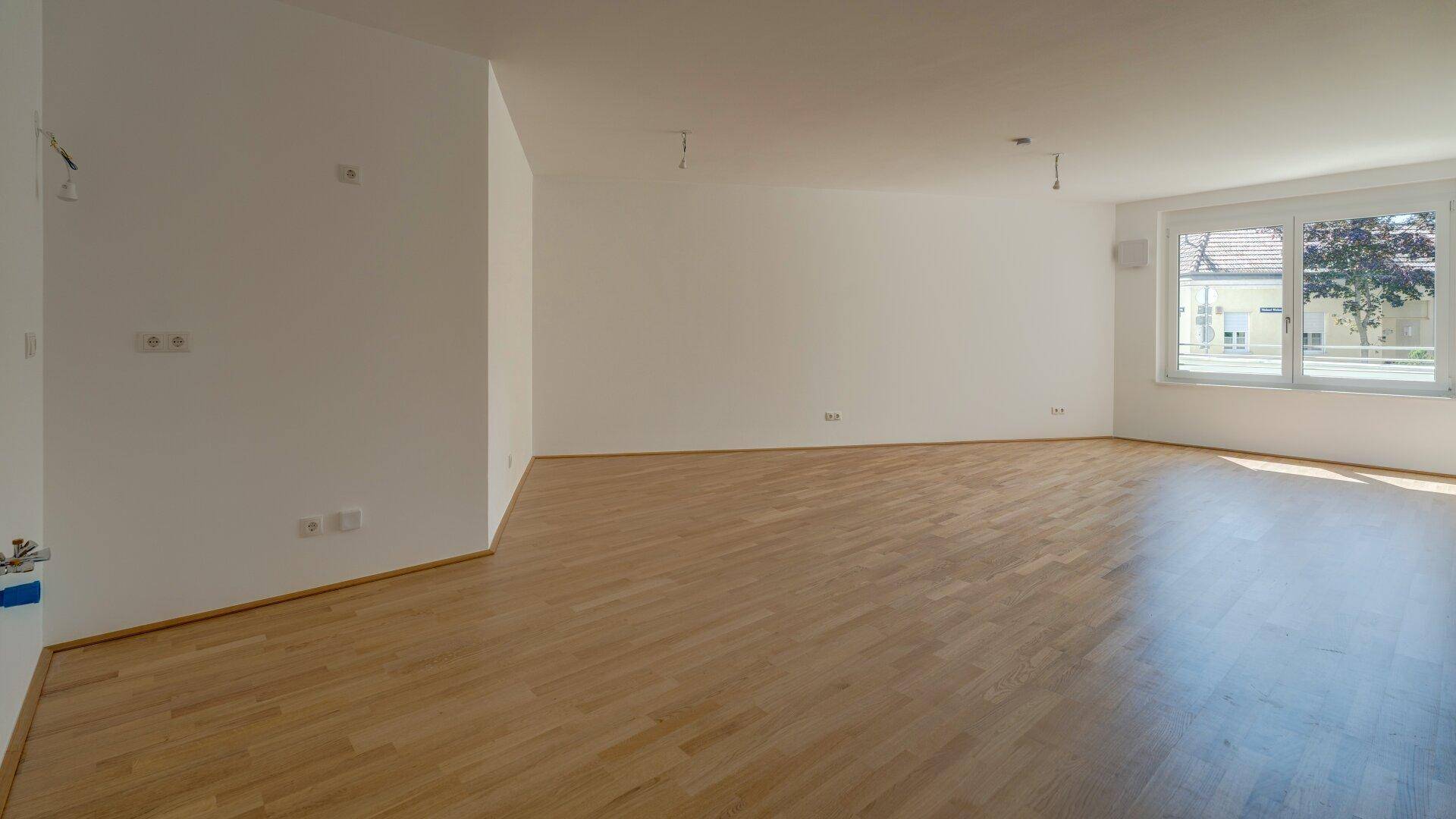 Geräumiges, leeres Wohnzimmer mit hellem Parkettboden und großen Fenstern für viel Tageslicht.