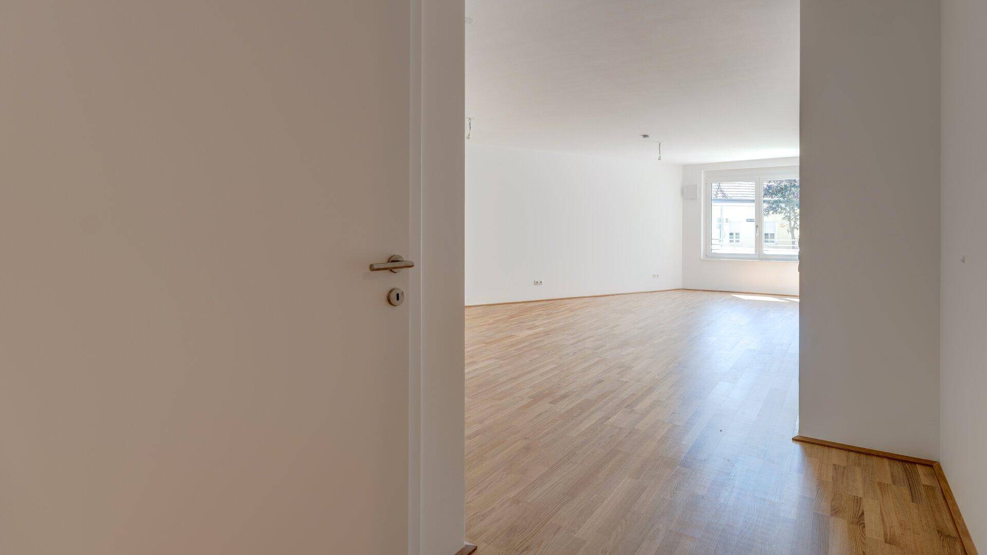 Blick durch eine offene Tür in ein helles Wohnzimmer mit Parkettboden und Tageslicht.