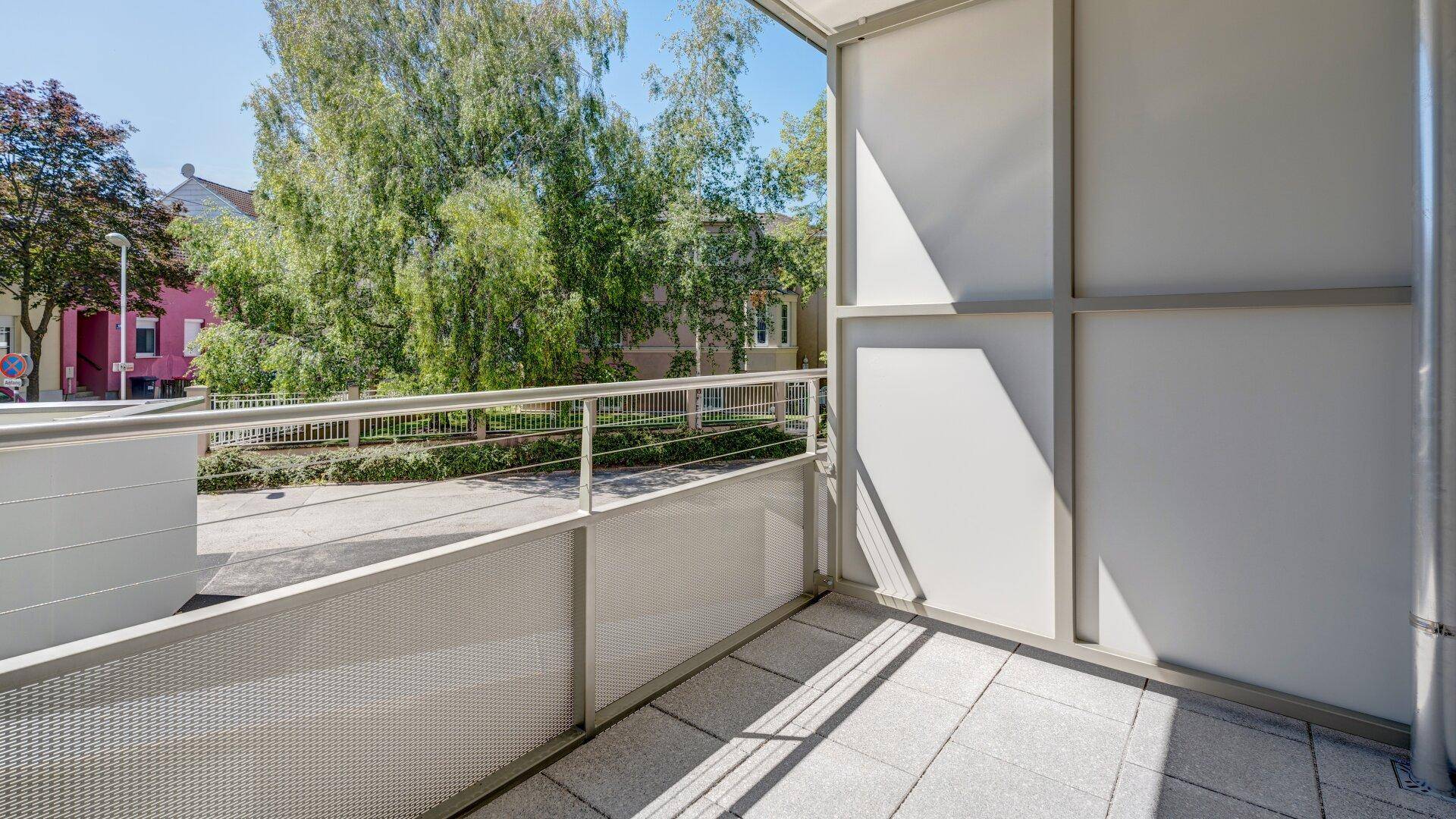 Heller Balkon mit Fliesenboden und Metallgeländer, umgeben von Bäumen und mit Blick auf die Straße.