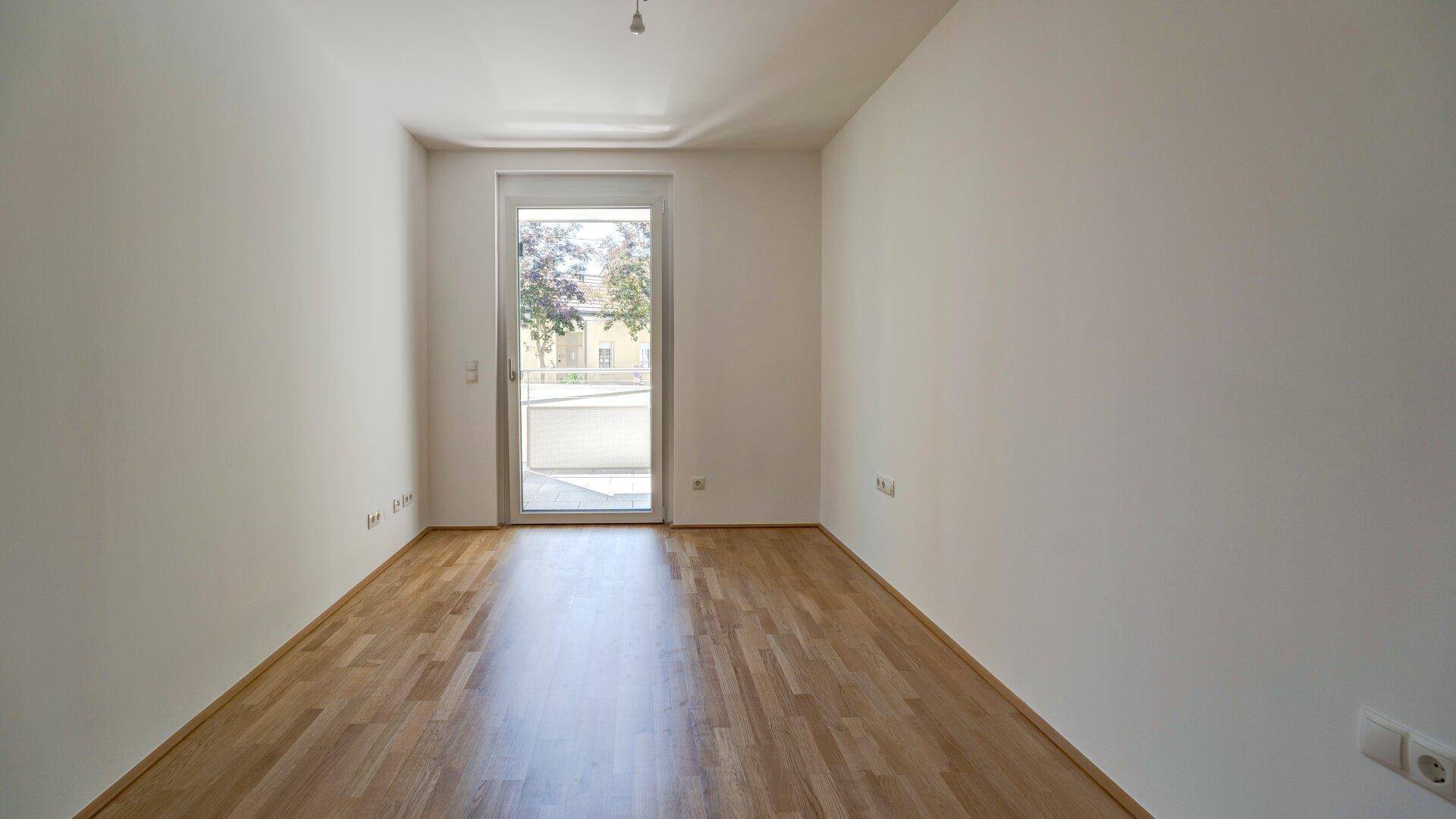 Leeres Zimmer mit Parkettboden und großer Balkontür, die viel Licht hereinlässt.