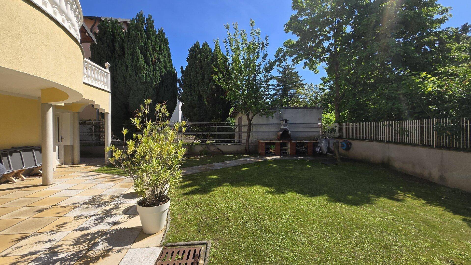 Großer, gepflegter Gartenbereich mit Rasenfläche, Bäumen und einem Grillplatz im Hintergrund.