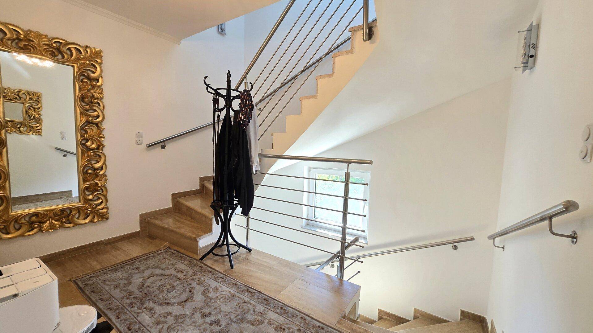 Heller Flur mit Holztreppe, modernem Geländer und einem großen, verzierten Spiegel.