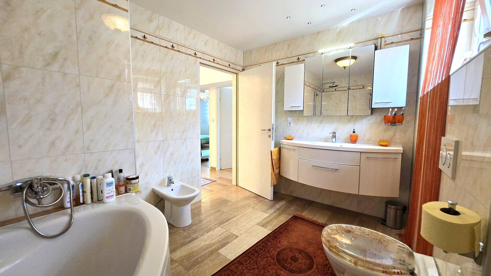 Helles Badezimmer mit Badewanne, Bidet, Doppelwaschbecken und Fliesen in Marmoroptik.