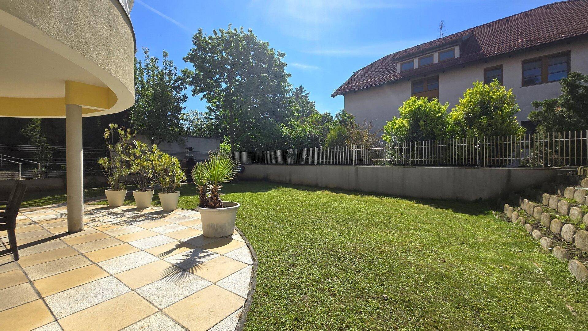 Sonniger Außenbereich mit gefliester Terrasse, Rasenfläche und Blick auf das umliegende Grün.