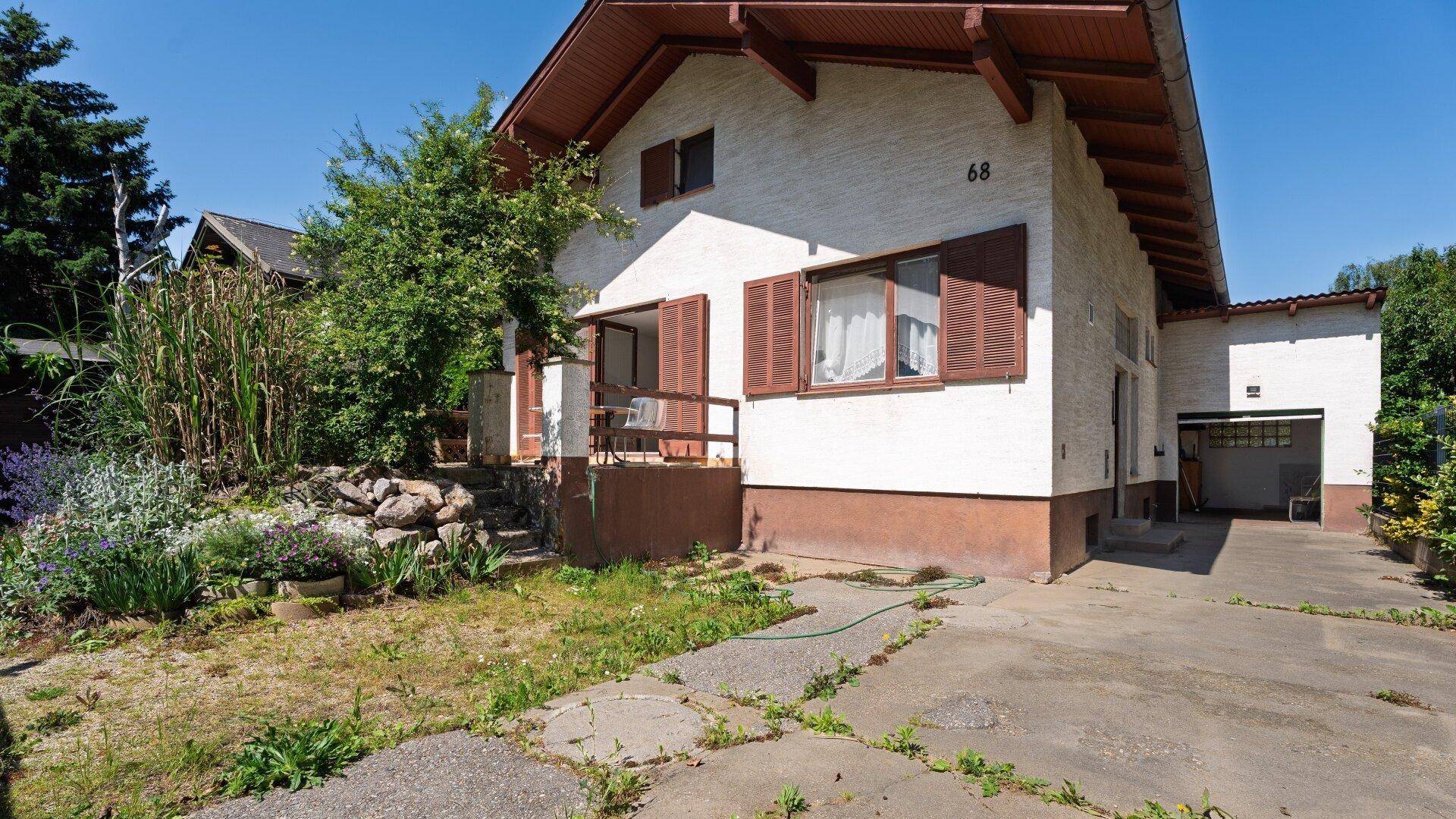 Vorderansicht des Hauses mit braunen Fensterläden, Einfahrt und Vorgarten an einem sonnigen Tag.