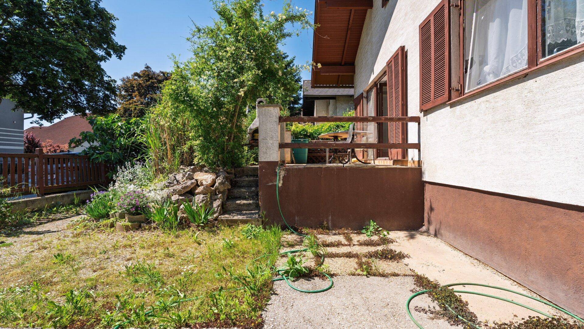 Terrasse am Haus mit Zugang zum Garten, umgeben von Pflanzen und mit Holzzaun.