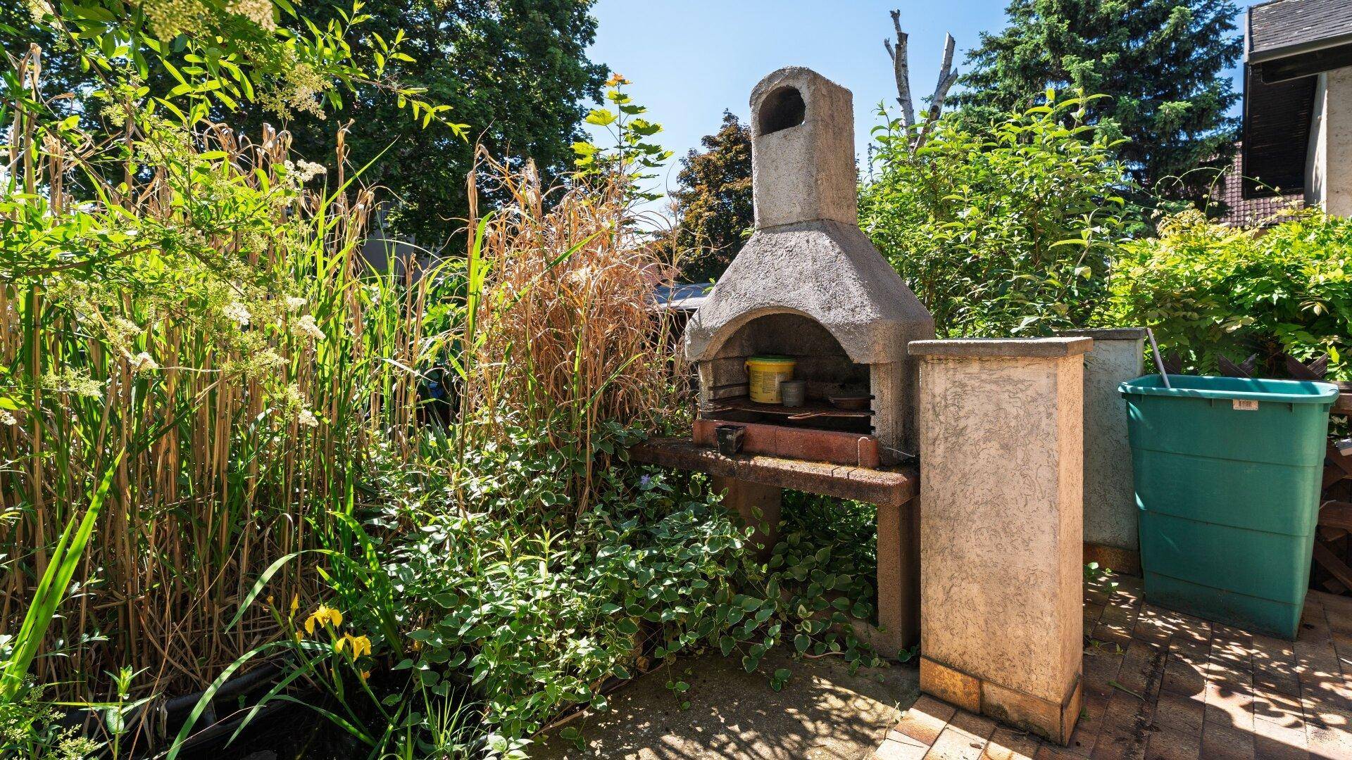 Gemauerter Grill im Garten, umgeben von üppiger Vegetation und hohen Gräsern.
