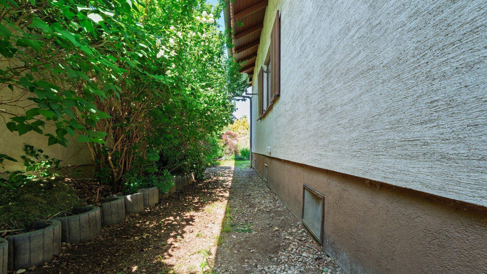 Schmaler Weg entlang der Hauswand, gesäumt von Büschen und kleinen Steinen.