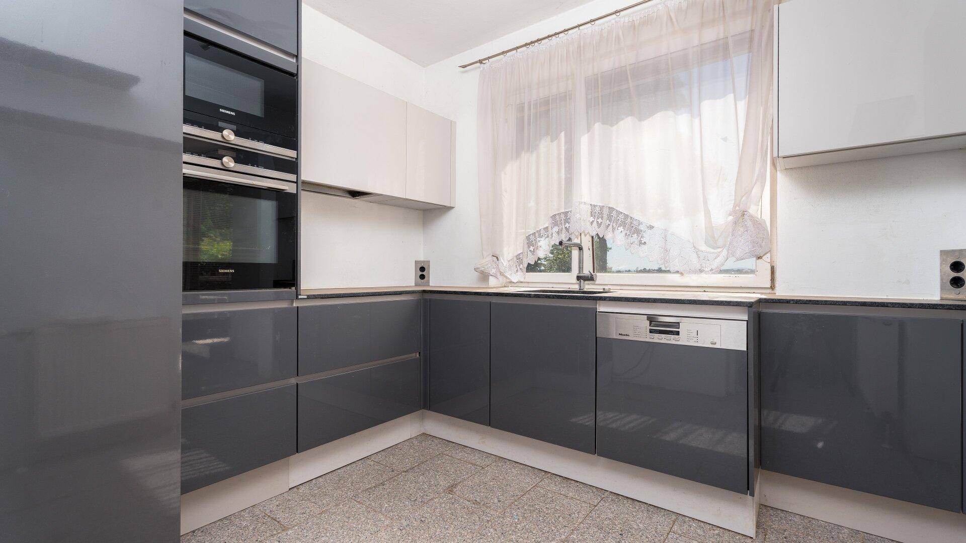 Moderne Küche mit grauen Hochglanzfronten, Einbaugeräten und Fenster mit Vorhang.