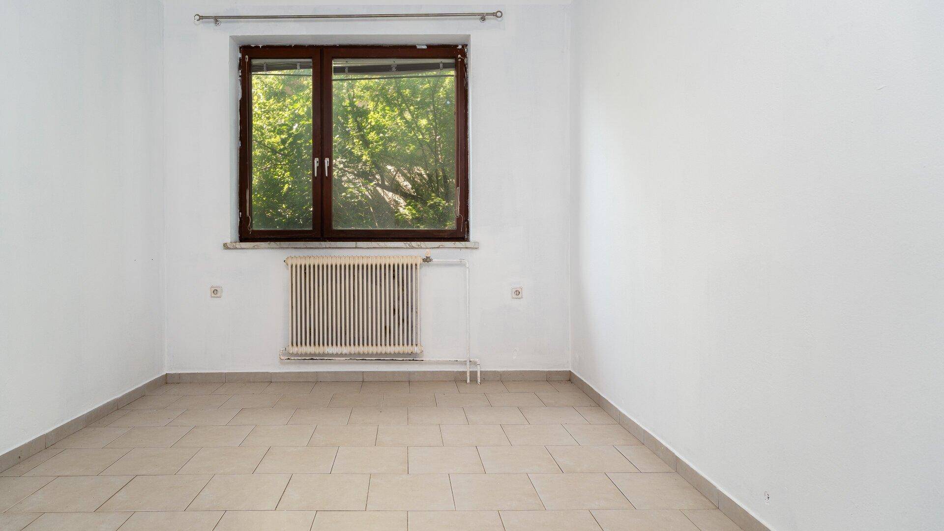 Helles Zimmer mit gefliestem Boden und großem Fenster mit Blick ins Grüne.