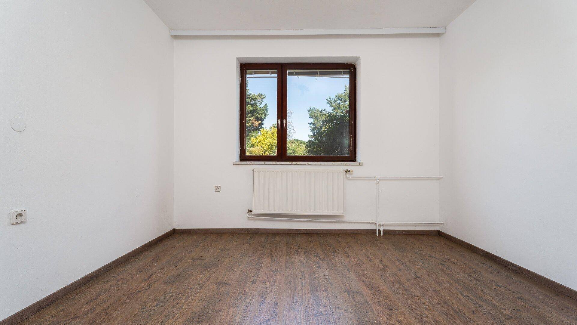 Leeres Zimmer mit Laminatboden und großem Fenster, das viel Tageslicht hereinlässt.