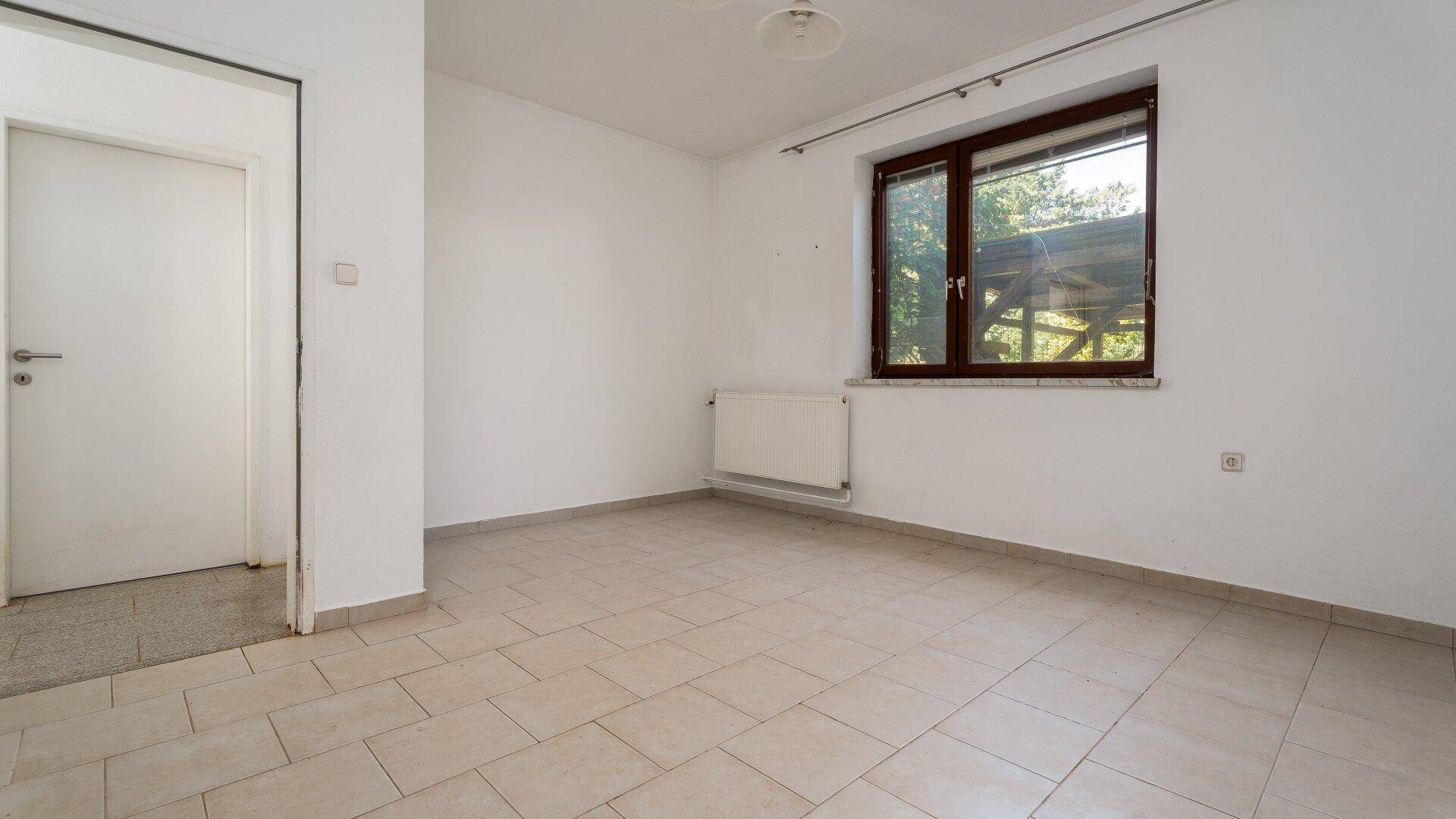 Helles Zimmer mit Fliesenboden und einem Fenster, das den Blick ins Grüne freigibt.