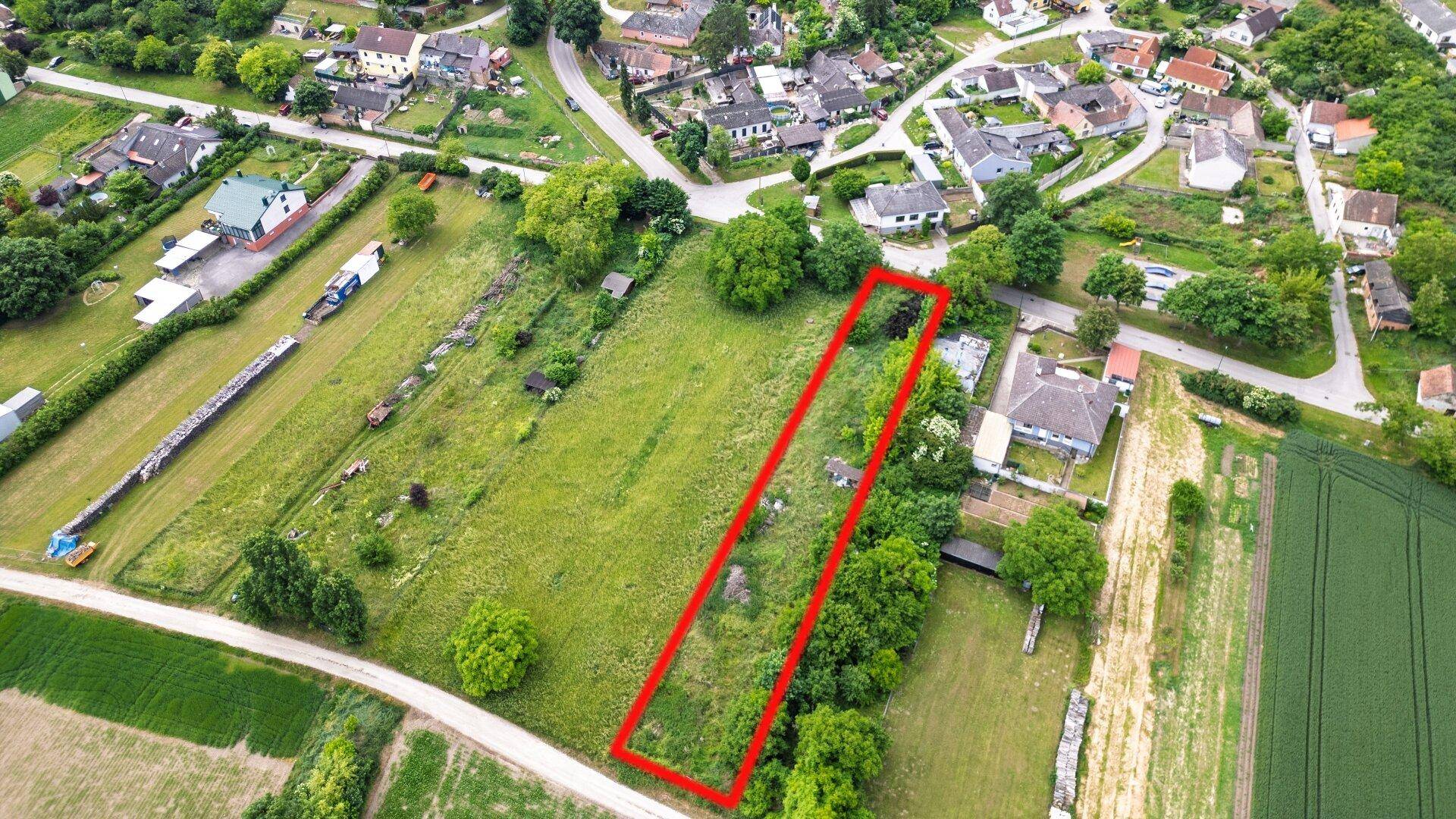 Schräge Luftaufnahme eines langgestreckten Grundstücks mit roter Umrandung in grüner Landschaft.