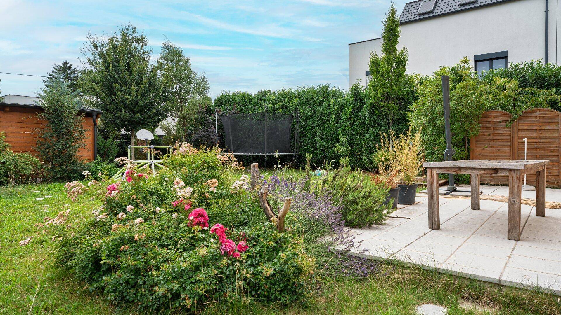 Gepflegter Garten mit Terrasse, Blumenbeeten, Rasenfläche und Trampolin.