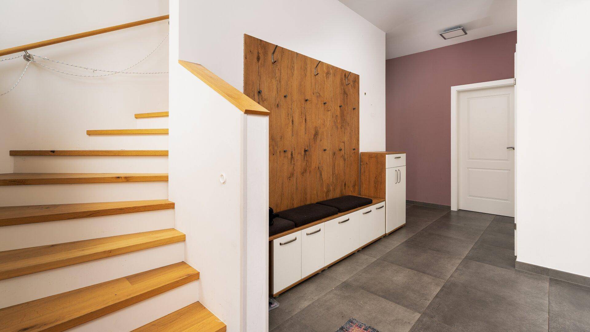 Moderner Flur mit Holztreppe, Garderobenbank und dunklen Bodenfliesen.