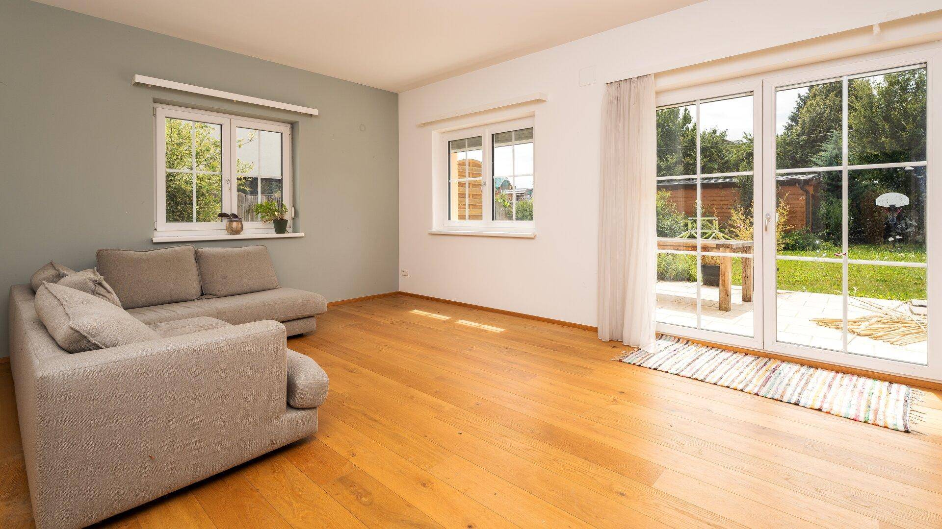 Helles Wohnzimmer mit Holzboden, Sofa und großen Fenstern zum Garten.