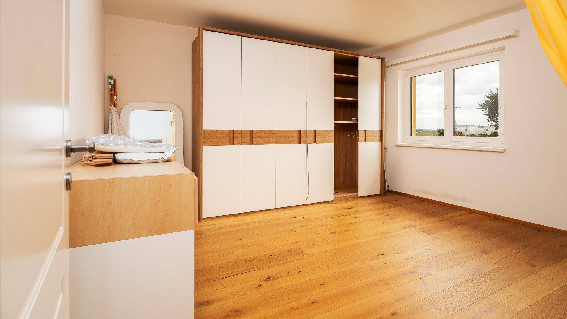 Helles Schlafzimmer mit Holzboden, Kleiderschrank und Blick ins Grüne.