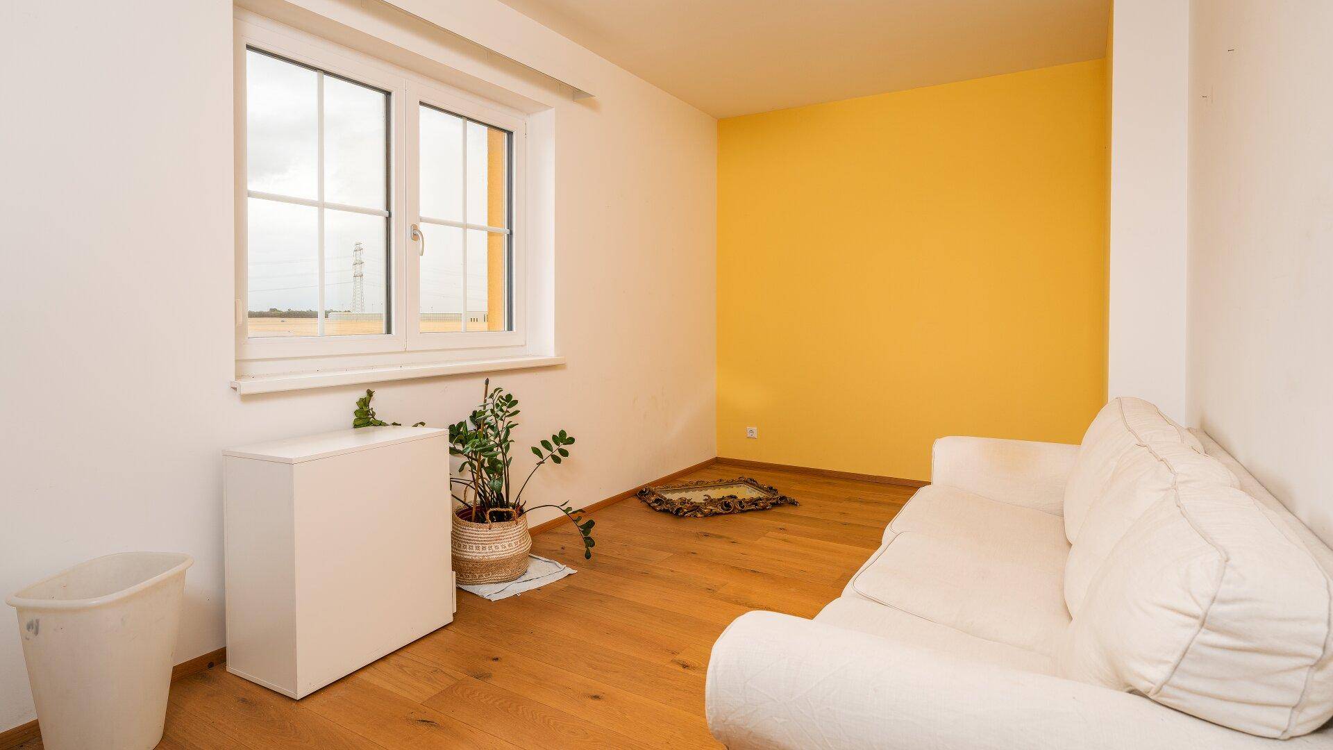 Helles Zimmer mit Holzboden, gelber Akzentwand und Blick ins Freie.