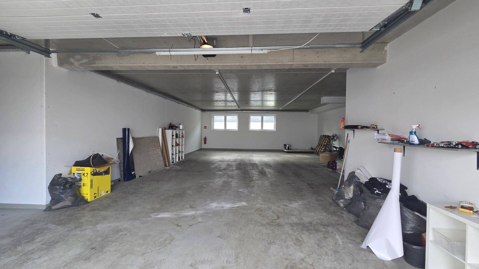 Weitläufiger Lagerraum mit Betonboden und hohen Decken, beleuchtet durch Fenster am Ende des Raumes.
