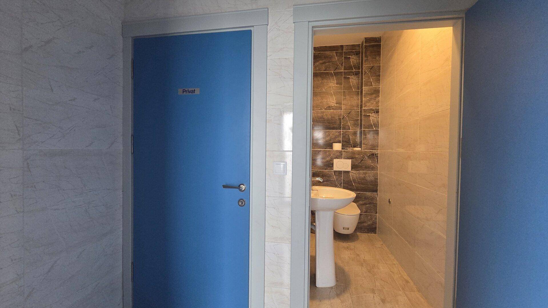 Eingangsbereich zu einem Badezimmer mit Waschbecken und Toilette, daneben eine geschlossene blaue Tür.