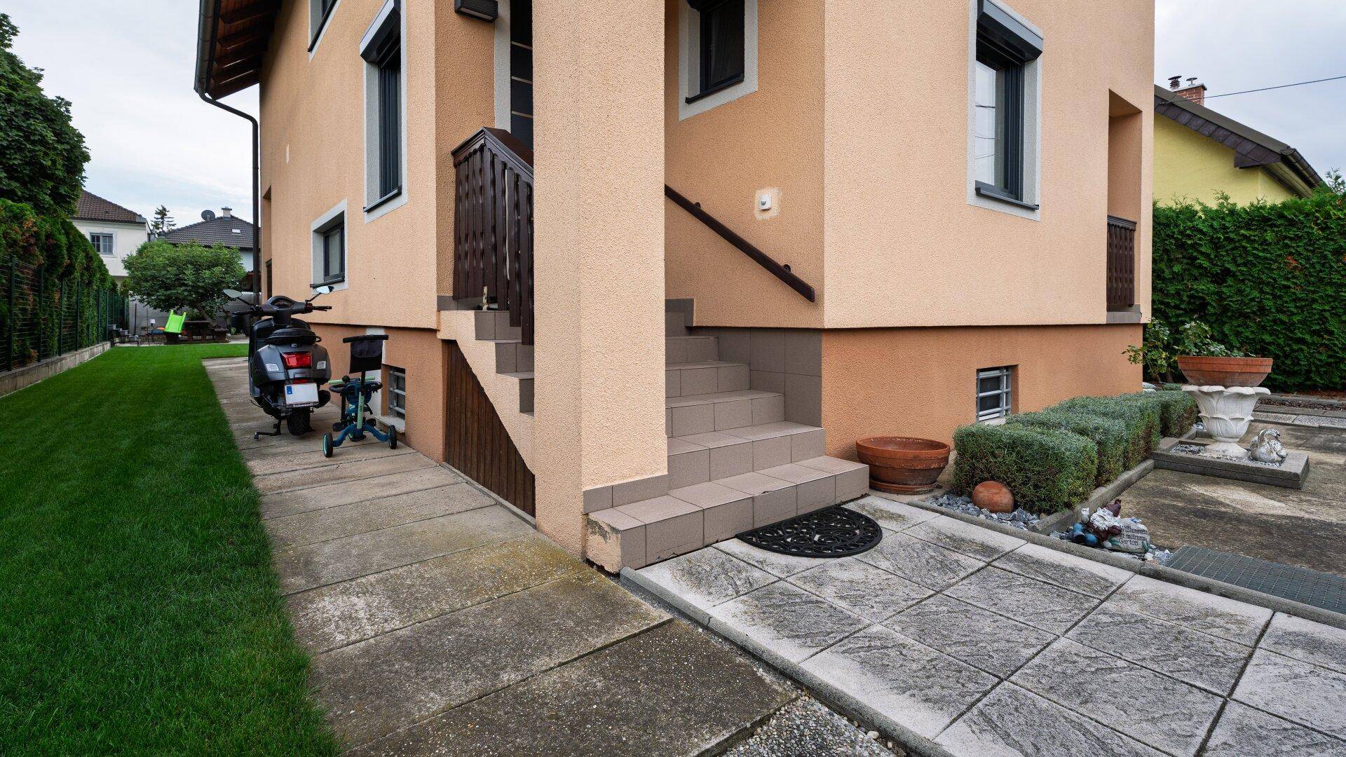 Detailansicht des Hauseingangs mit Treppe und Zugang zum Untergeschoss, umgeben von kleinen Büschen.