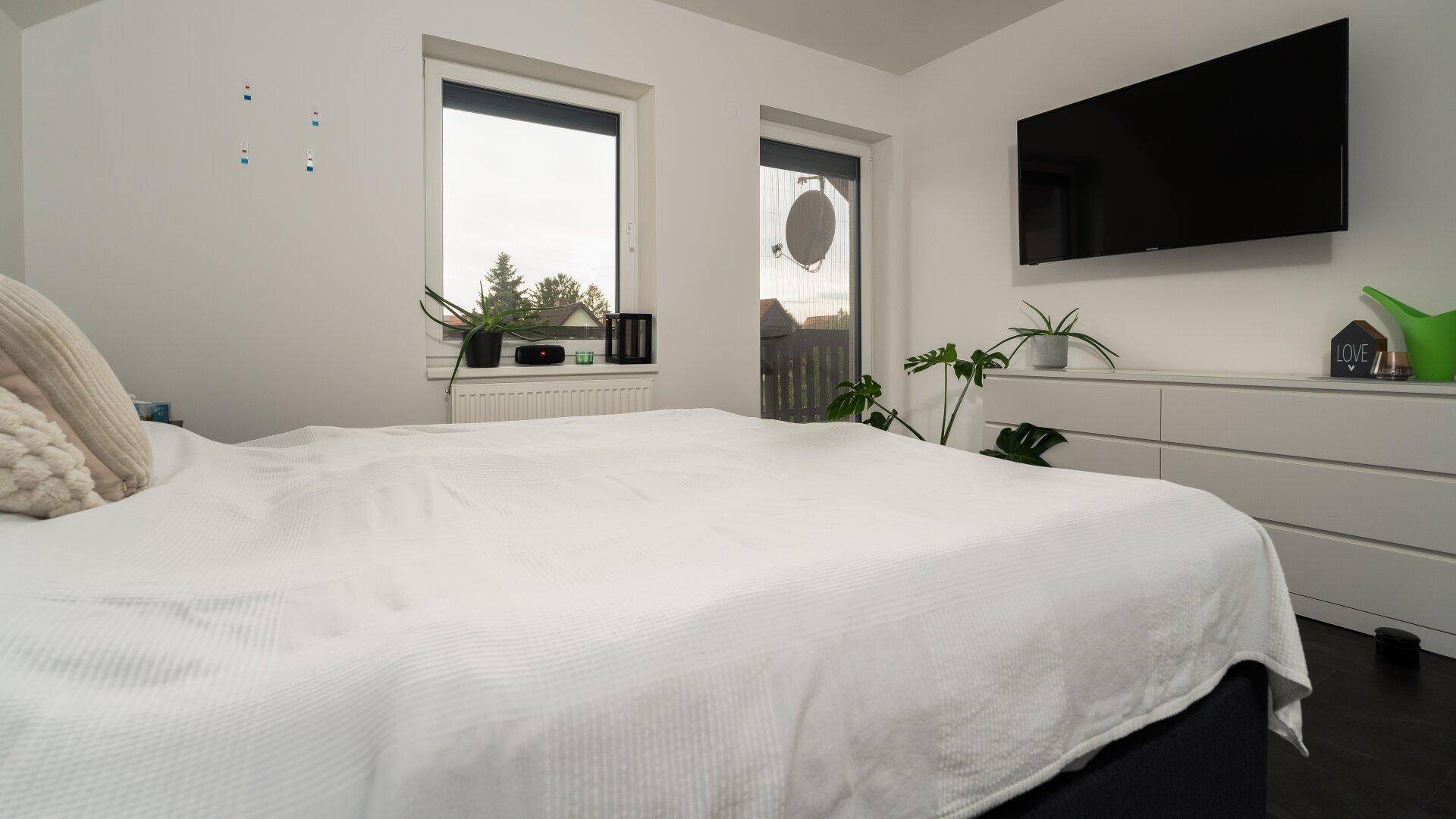 Helles Schlafzimmer mit großem Bett, Kommode und Blick ins Grüne durch die Fenster.