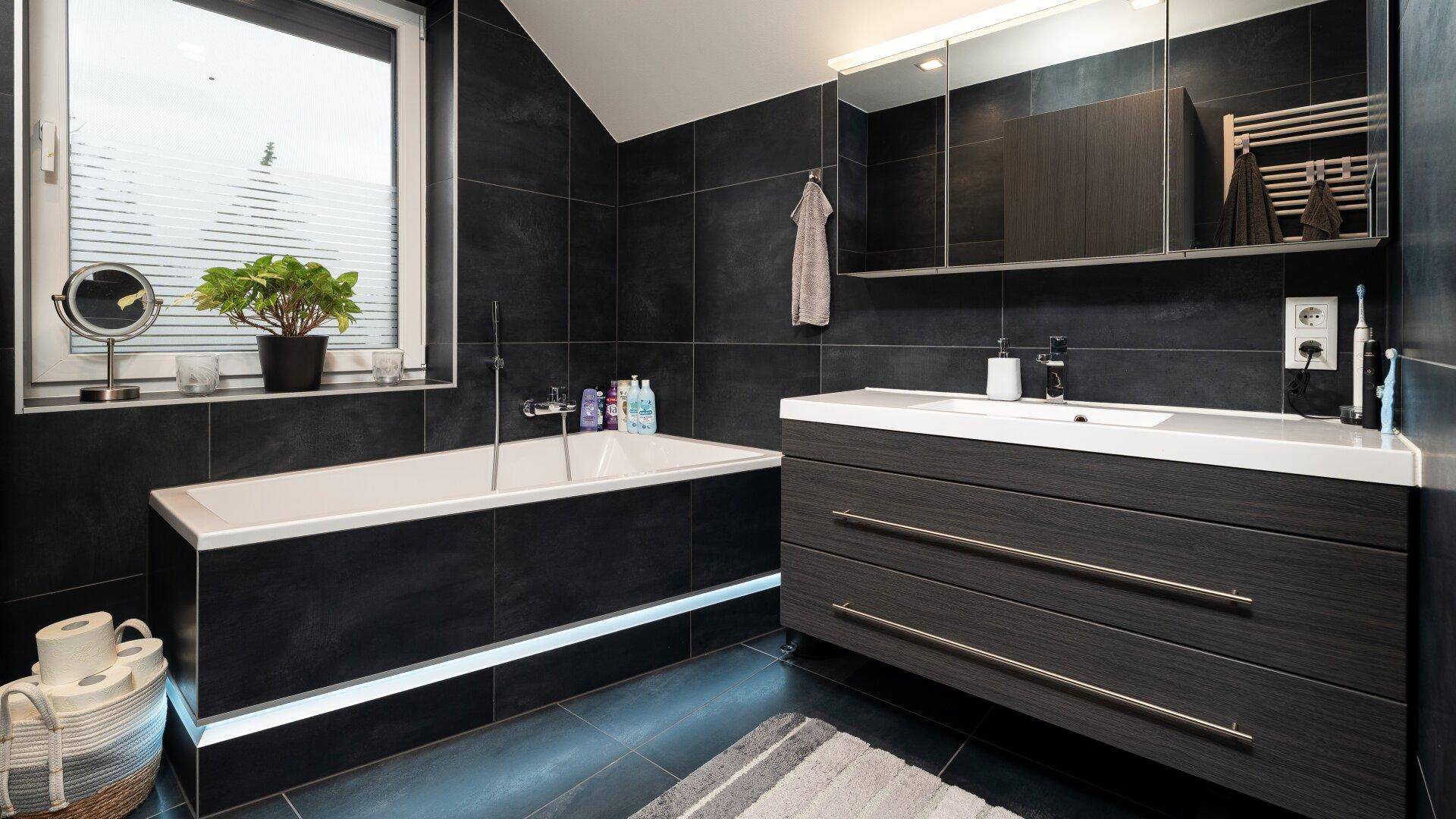 Modernes Badezimmer mit dunklen Fliesen, Badewanne, Waschtisch und Spiegelschrank.