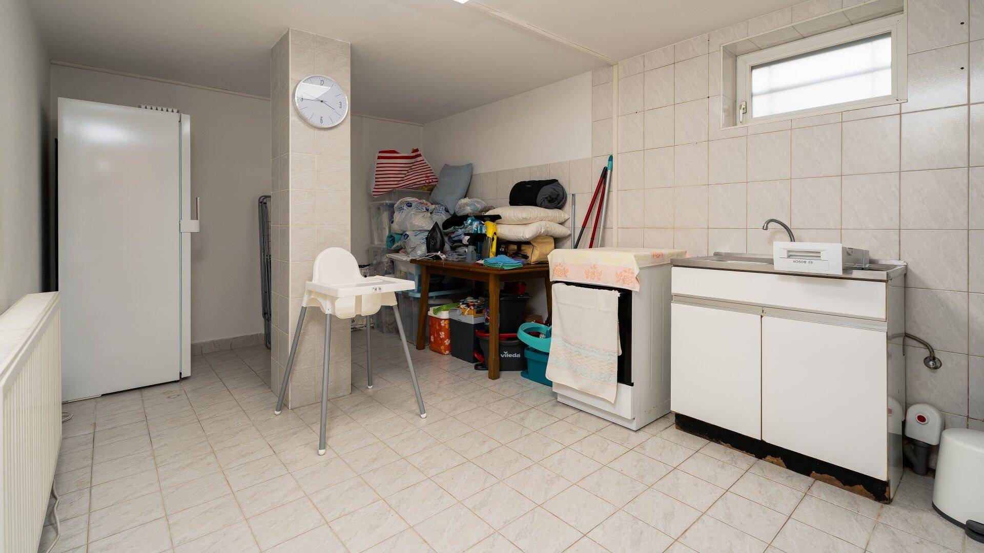 Gefliester Hauswirtschaftsraum im Keller mit Waschbecken, Kühlschrank und kleinem Fenster.