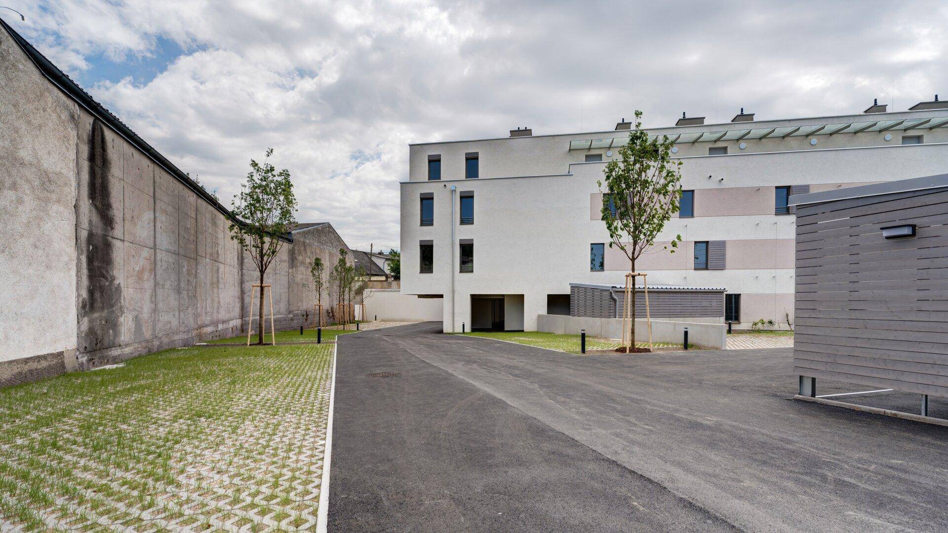 Außenansicht der modernen Wohnanlage mit Zufahrt und angrenzender alter Mauer unter bewölktem Himmel.