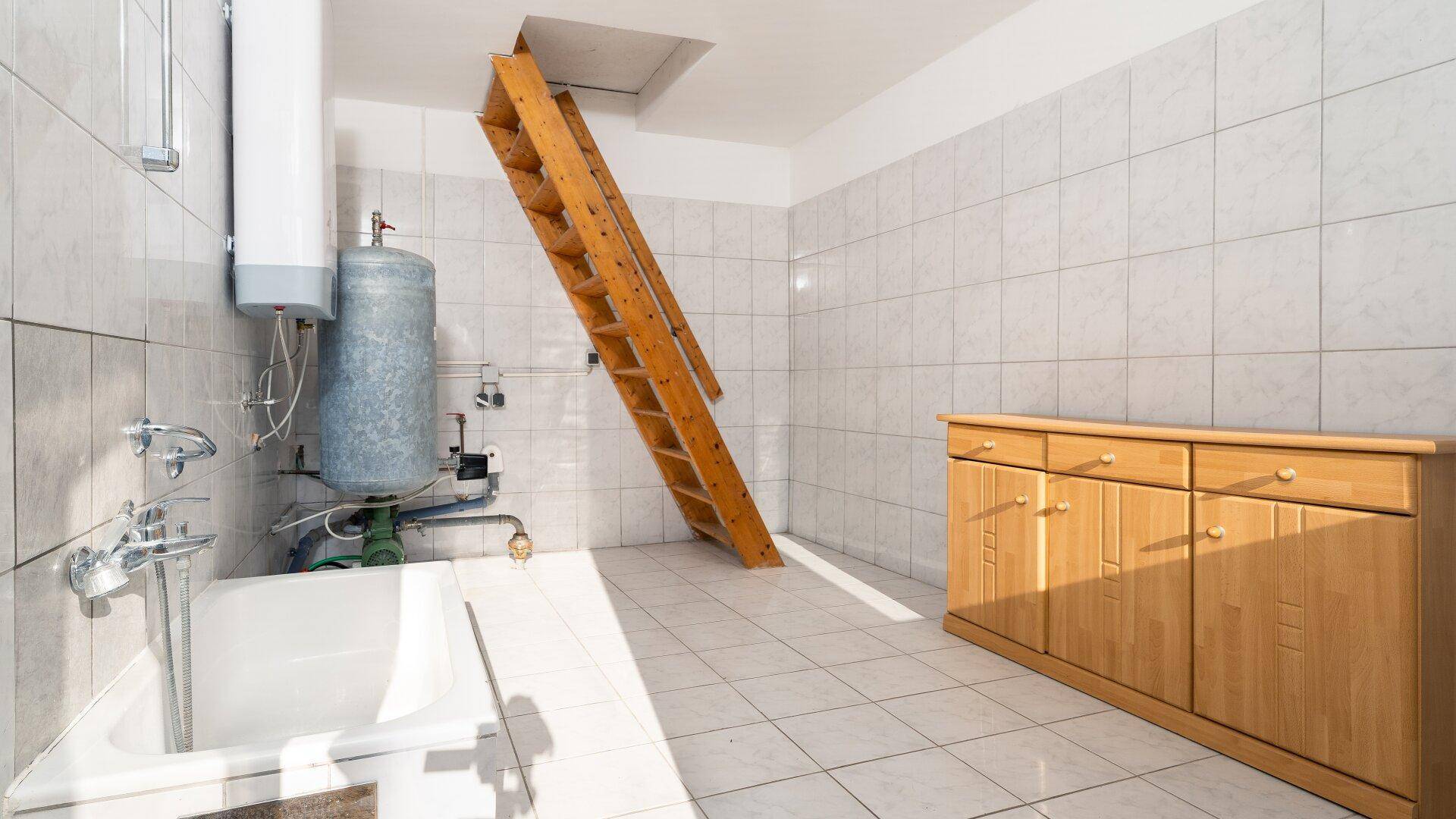 Hauswirtschaftsraum mit Badewanne, Warmwasserboiler und Zugang zum Dachboden über eine Leiter.