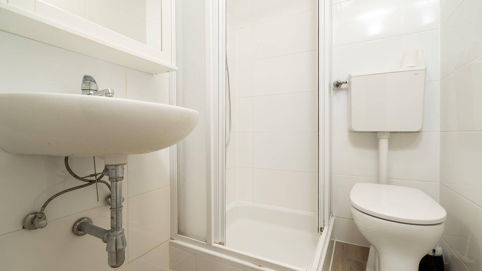 Kleines Badezimmer mit WC, Waschbecken und Duschkabine, komplett gefliest in Weiß.