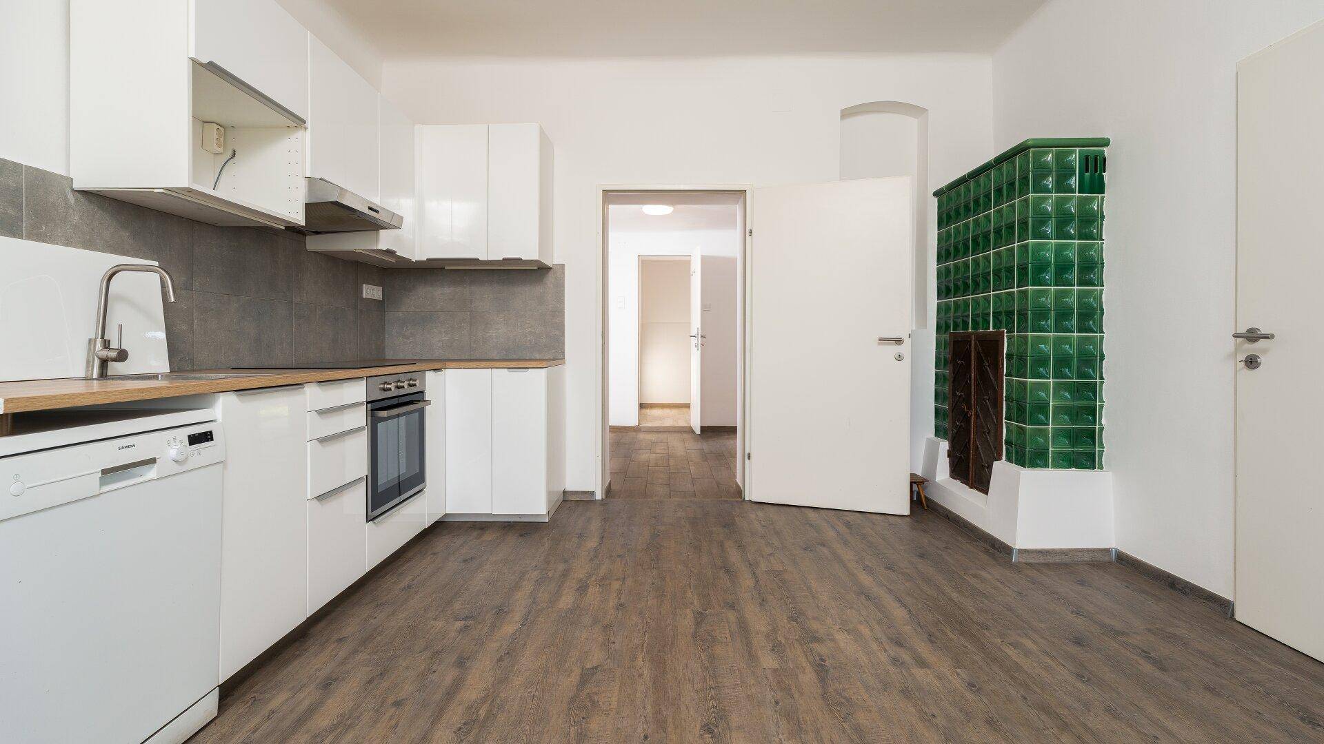Offene Küche mit weißer Einrichtung, Laminatboden und einem grünen Kachelofen.