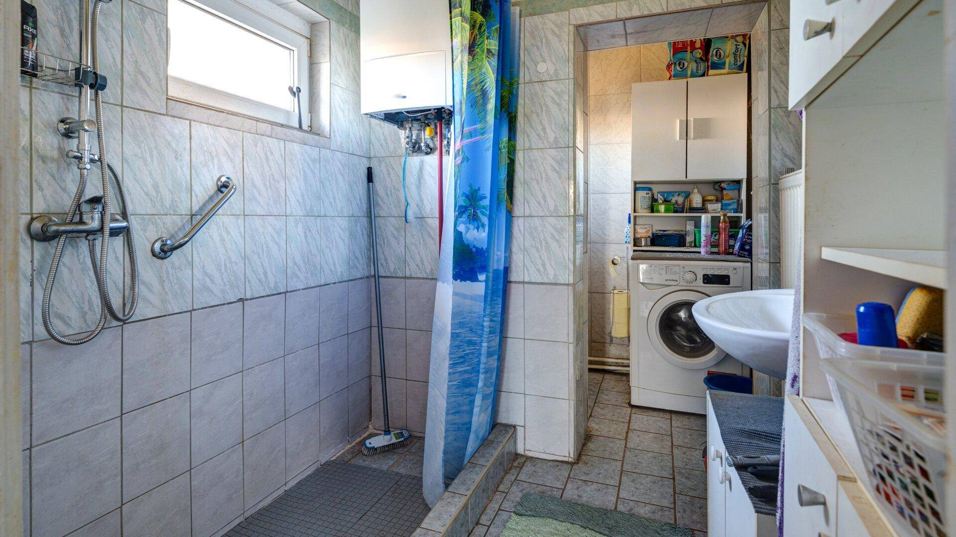 Gefliestes Badezimmer mit Duschbereich, Waschmaschine und Haltegriffen für Barrierefreiheit.