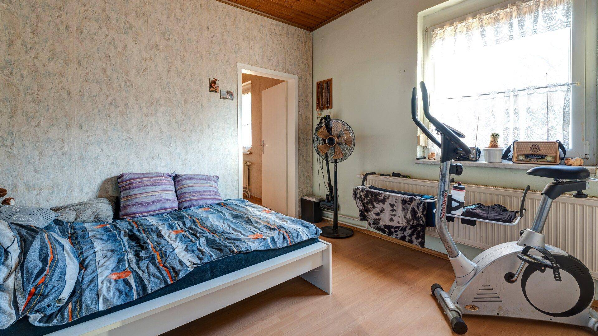 Schlafzimmer mit Bett, Heimtrainer und einem Fenster, das viel Tageslicht hereinlässt.