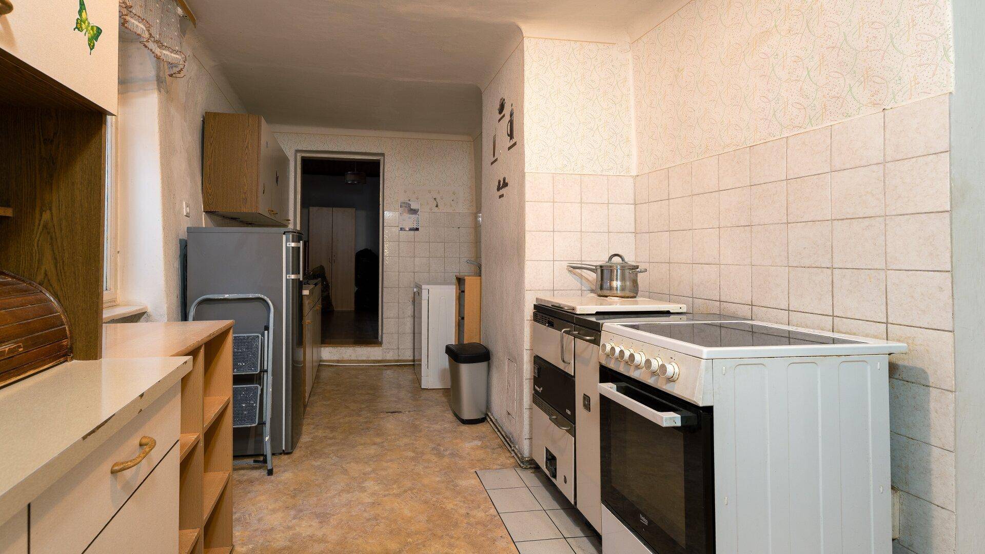 Einfache Küche mit älteren Geräten, Fliesenwand und Blick in einen weiteren Raum, funktional eingerichtet.