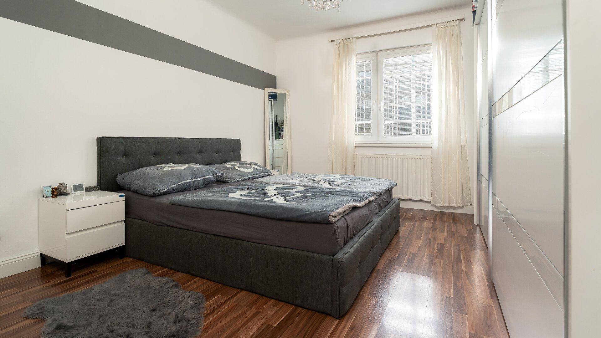 Schlafzimmer mit Holzboden, großem Bett und weißem Kleiderschrank, gut beleuchtet durch ein Fenster.